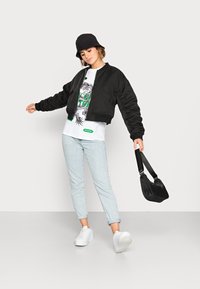Casaco bomber preto cropped com uma textura suave, combinado com uma t-shirt branca com gráfico, jeans de ganga claros e ténis brancos. Chapéu de balde preto.