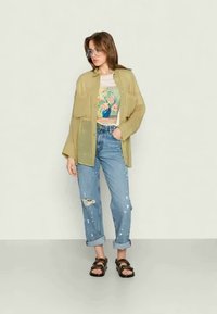 Camisa leve, verde oliva, sobre um top de flores, combinada com jeans claros destruídos e sandálias castanhas escuras.