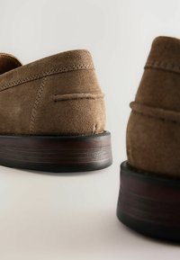 Mocasines de ante marrón con un perfil bajo y una suela de goma negra texturizada. Detalles de costura a lo largo de los lados y la parte posterior.