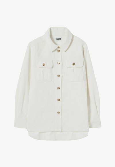 Camisa de corduroy blanca con mangas largas, cuello puntiagudo, cierre frontal de botones, dos bolsillos en el pecho y botones en tono dorado.