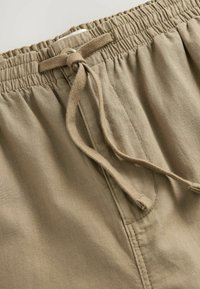 Khaki sort elasztikus derékkal, szövet zsinórral és sima felülettel. Csomózó részletek és egy standard zseb jellemzi.