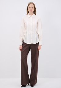 Crème kleurig shirt met lange mouwen en een knopenfront, voorzien van verticale plooien en een klassieke kraag, gecombineerd met hoge bruine broeken.