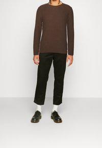 Chemise marron côtelée à manches longues associée à un pantalon noir fuselé et à des chaussures noires à lacets. Des chaussettes blanches visibles au-dessus des chaussures.