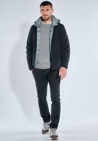 Black padded jacket with gray inner lining, ribbed gray sweater, dark pants, and beige sneakers. Model stands against a white background.