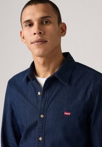 Tamno plavi traper košulja na dugme s klasičnim ovratnikom, bež dugmadi i crvenim Levi's logotipom na lijevoj prednjoj džepu.