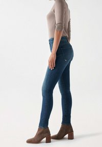 Jeans ajustados de mezclilla en color azul con cintura alta, combinados con botines marrones de tobillo. La parte de arriba es un suéter ceñido de color beige claro.