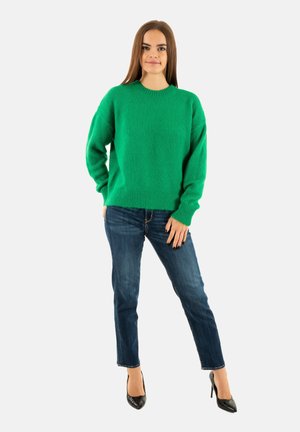 Pull en maille verte avec un col rond et une coupe décontractée, associé à un jean bleu foncé et des escarpins noirs à bout pointu. Texture lisse, design décontracté.