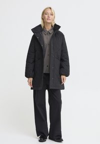 Zwarte gewatteerde jas met een grote kraag, knoopsluiting en twee voorkruiden. Gedragen over een grijze knoopblouse en wijde broek.