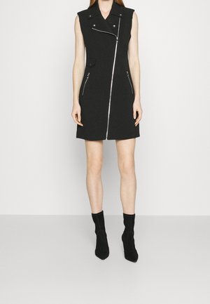 Femme portant une mini-robe noire sans manches avec une fermeture éclair asymétrique à l'avant, des poches zippées et des bottines noires.