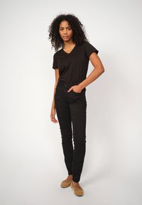 Čierne tričko s výstrihom do V a čierne slim-fit nohavice vyrobené z jemnej látky. Model má na sebe béžové sklápacie topánky a stojí na svetlom pozadí.