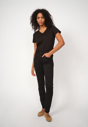 Černé tričko s véčkovým výstřihem a černé slim-fit kalhoty, vyrobené z měkké látky. Model má na sobě béžové slip-on baleríny, stojí proti světlému pozadí.