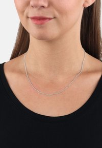 Srebrny naszyjnik o gładkim, smukłym kształcie spoczywający na obojczyku. Noszony na czarnej bluzce, podkreślający minimalistyczny styl.