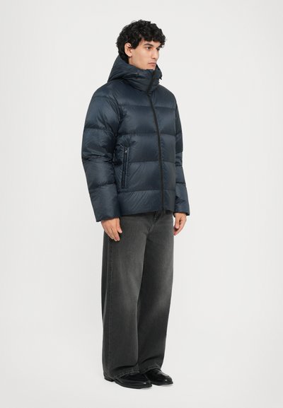 Marineblauer Pufferjacke mit hohem Kragen, Reißverschluss und zwei Seitentaschen; kombiniert mit lässigen grauen Jeans und schwarzen Schuhen.