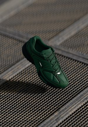 Baskets de sport vert foncé avec semelle noire et logo argenté, flottant au-dessus d'un fond de grille métallique texturée.