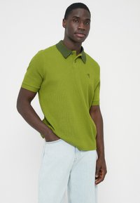 Élénk zöld színű galléros póló sötétebb zöld gallérral. Rövid ujjú, texturált anyagból, bal oldalon a mellrészen egy kis logóval. Világos farmerrel párosítva.