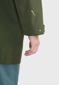 Manica di giacca impermeabile verde con un pulsante a pressione argento e un design logo sottile vicino al polsino. Tessuto dalla texture liscia.