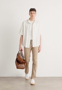 Wit gestreept shirt met korte mouwen, crèmekleurig T-shirt, beige broek en witte sneakers. Bruin leren sporttas met handvatten.