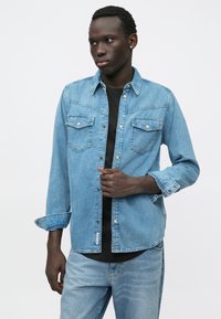 Νεαρός άνδρας φορώντας ανοιχτό μπλε τζιν μπουφάν και τζιν με μαύρο πουκάμισο, στέκεται μπροστά σε λευκό φόντο, κοιτάζοντας στο πλάι.