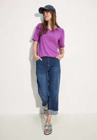 T-shirt violet à manches courtes en V, jeans bleu à jambes larges avec ourlets retournés, et sandales argentées à deux brides avec boucles. Tenue décontractée.