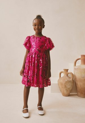 Vestido de lentejuelas fucsia con mangas cortas abullonadas y cintura ajustada, adornado con patrones florales. Combinado con zapatos Mary Jane blancos.