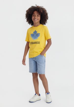 Χαμογελαστό παιδί με σγουρά μαλλιά που φοράει κίτρινη μπλούζα DSQUARED2, ανοιχτόχρωμα τζιν σορτς και λευκά αθλητικά παπούτσια, στέκεται με το ένα χέρι στην τσέπη.