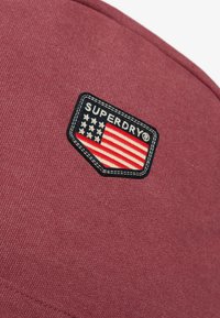 Parche de tela color marrón con el diseño de "SUPERDRY" y una bandera americana. Presenta bordado y costura en el borde para un detalle adicional.