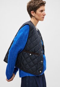 Fekete steppelt mellény patentos gombokkal, kék texturált pulóver fölött viselve. Barna díszzsebbel rendelkezik. A modell oldalról áll.