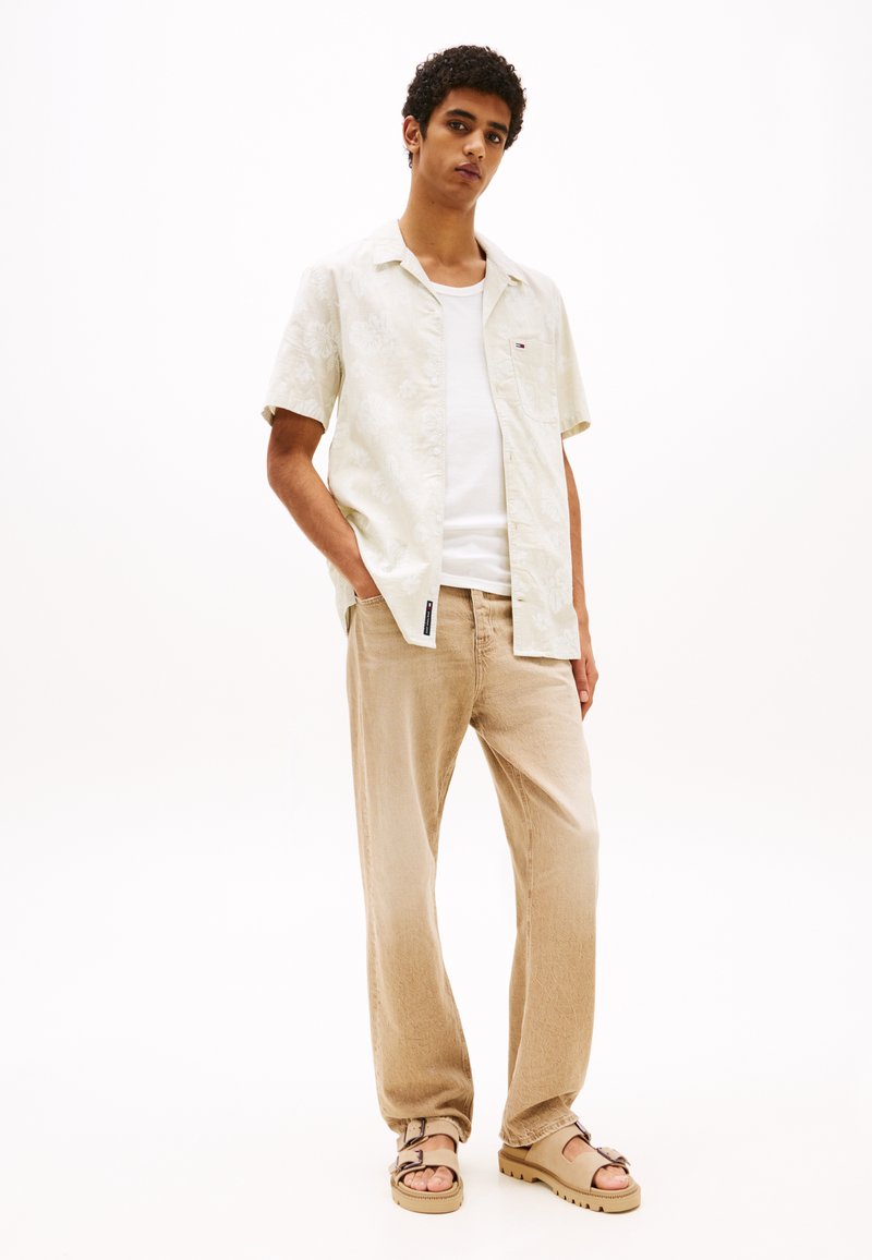 Chemise à manches courtes légère à motifs floraux, pantalon en velours côtelé beige, sandales marron avec boucle, portées avec un débardeur blanc, fond neutre.