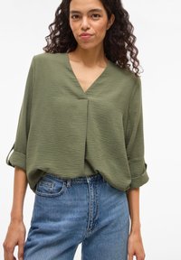 Πράσινο μπλουζάκι με υφή, V-λαιμόκοψη, μανίκια τριών τετάρτων και άνετη εφαρμογή, σε συνδυασμό με ανοιχτό μπλε ψηλόμεσα τζιν.