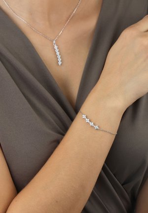 Silver necklace with a linear pendant featuring several connected clear gemstones; matching bracelet with similar gemstone accents, worn on skin.