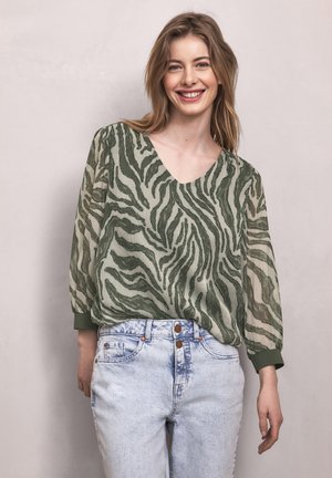 Uśmiechnięta kobieta o jasnobrązowych włosach, ubrana w zieloną prześwitującą bluzkę w zebrowy wzór oraz jasnoniebieskie, wysokowalne jeansy, na jednolitym tle.