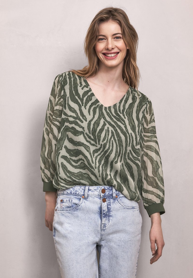 Femme souriante aux cheveux châtain clair portant une blouse transparente à rayures zèbre vertes et un jean taille haute bleu clair sur un fond uni.