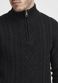Schwarzer Reißverschluss-Pullover mit einem hohen gerippten Kragen und einem Zopfmuster-Design, das vertikale Strukturen über den Stoff zieht.