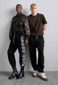Schwarze, taillierte Lederjacke, kombiniert mit schwarzen Jogginghosen, die weißen Text haben. Das Modell trägt elegante schwarze Stiefel, während das männliche Modell ein braunes T-Shirt und klobige Sneaker trägt.