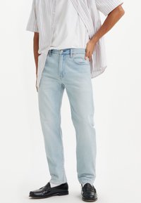 Jeans de mezclilla azul claro con corte recto, llevados con una camiseta blanca y una camisa de manga corta a rayas multicolores, acompañados de mocasines negros.