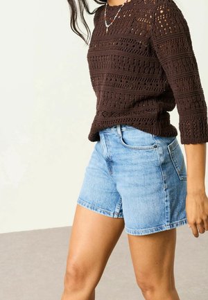 Personne portant un pull en maille crochet brun foncé à manches trois-quarts et un short en jean bleu clair, debout devant un fond neutre.