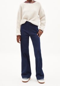Maglione crema a coste con un collo multicolore e testurizzato, abbinato a jeans a zampa d'elefante blu scuro e sneakers grigio chiaro con suole marroni.