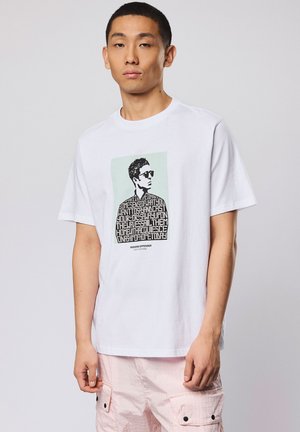 Junger Mann trägt ein weißes T-Shirt mit schwarz-weißem Porträt und Text, kombiniert mit hellrosa Cargo-Shorts vor neutralem Hintergrund.