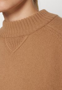 Pull marron tricoté avec un col montant côtelé, présentant une texture lisse et un détail discret de couture triangulaire.