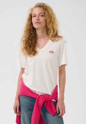 Femme aux cheveux blonds bouclés portant un t-shirt blanc à col en V avec un emblème de lèvres rouges et un pull rose noué autour de la taille.