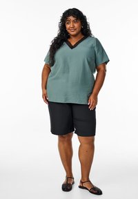 Top corto e ampio con maniche corte in tessuto verde testurizzato, con scollatura in pizzo nero; abbinato a pantaloni corti neri fino al ginocchio e sandali neri.