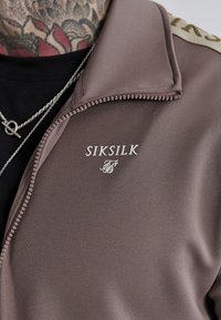 Chaqueta marrón con cremallera de textura suave; presenta un logo de marca "SIKSILK" en blanco y un collar con un detalle en contraste.