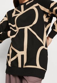 Czarny sweter z beżowymi, abstrakcyjnymi wzorami geometrycznymi, noszony przez osobę z rękami na biodrach, zestawiony z malinowymi spodniami.