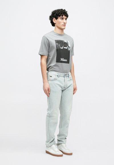 Γκρι βαμβακερό μπλουζάκι με μαύρο και άσπρο γραφικό σχέδιο και το κείμενο "Milano". Ανοιχτό μπλε τζιν παντελόνι και λευκά αθλητικά παπούτσια ολοκληρώνουν το σύνολο.