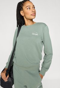 Lysegrønn genser med ribbede mansjetter og kant, med hvit "LIMITLESS"-tekst. Modellen bærer en svart veske og står foran en ensfarget bakgrunn.