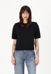 Schwarzes kurzärmliges Strickoberteil mit rundem Ausschnitt, kombiniert mit hellblauen Jeans, die an der Vorderseite mit Verzierungen ausgestattet sind.