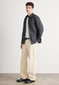 Camicetta button-up grigia scura sopra un maglione bianco, abbinata a pantaloni beige a gamba larga e scarpe nere. Tessuto morbido con una vestibilità rilassata.