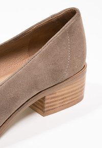 Chaussure à talons blocs en suede taupe avec un bout arrondi et un talon en bois. Doublure intérieure lisse et détails de couture sur la tige.