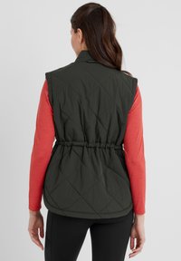 Quilted dark green vest with a fitted waist and snap collar, worn over a red long-sleeve shirt. Features seamless stitching and soft texture.