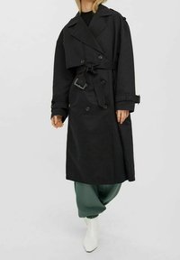 Czarny płaszcz trench z podwójnym zapięciem, szerokimi klapami, detalem w postaci pasa i zapinanymi mankietami, połączony z zielonymi spodniami i białymi butami.